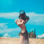 A hand raises a camera amidst a sunny field, evoking creativity and leisure.